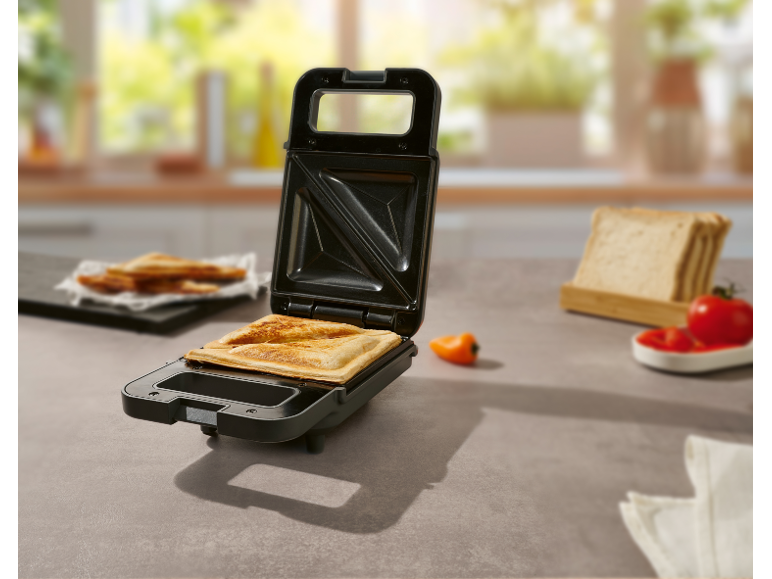 Tostapane aperto con un toast in cucina, con pane e pomodori sullo sfondo.