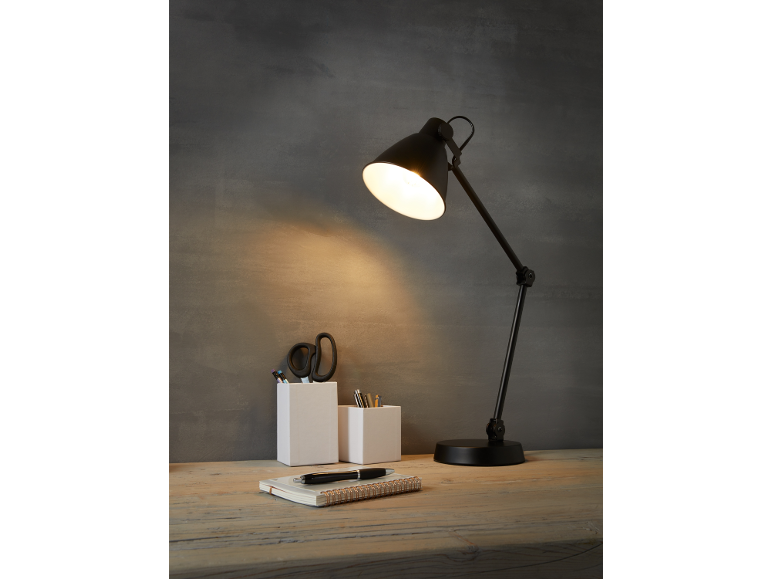 Lampada da scrivania nera illumina una scrivania in legno con un quaderno e penne in contenitori bianchi.