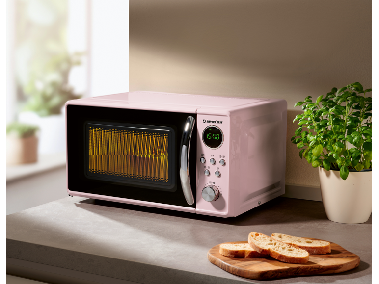 Microonde rosa su un bancone da cucina con erbe aromatiche e fette di pane.