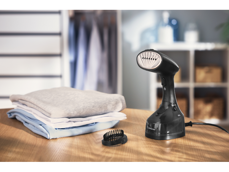 Vaporizzatore per indumenti nero con accessori e vestiti piegati su un tavolo di legno.