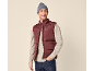 Uomo sorridente con gilet imbottito bordeaux, maglione beige e berretto grigio.