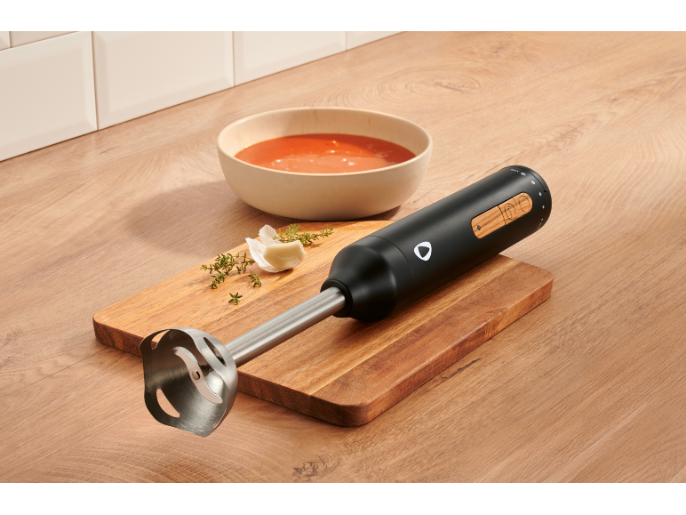 Frullatore a immersione nero con dettagli in legno, ciotola di zuppa di pomodoro ed erbe su tavolo di legno.