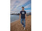 Uomo in t-shirt blu navy con stampa 'Original Quality' e jeans che cammina su una spiaggia sabbiosa.