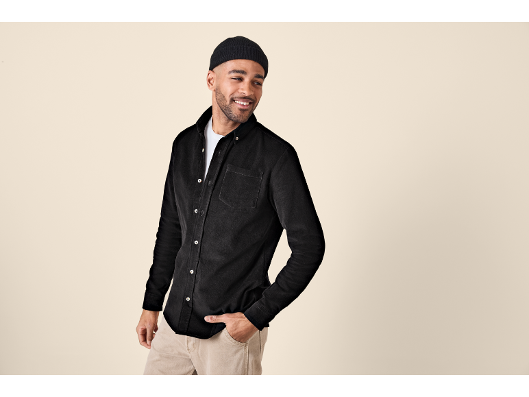 Uomo con camicia di velluto a coste nera e pantaloni beige, sorridente e che guarda altrove.