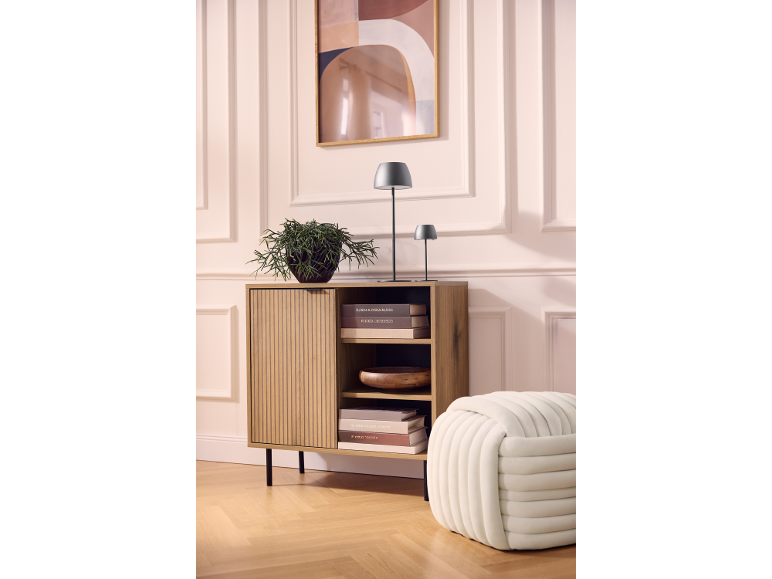 Mobile in legno con pianta, lampade e libri, accanto a un pouf, contro una parete con quadro.