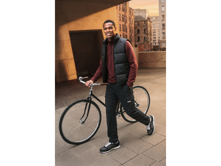 Uomo con gilet imbottito nero, maglia rossa a maniche lunghe e pantaloni cargo neri con bicicletta.