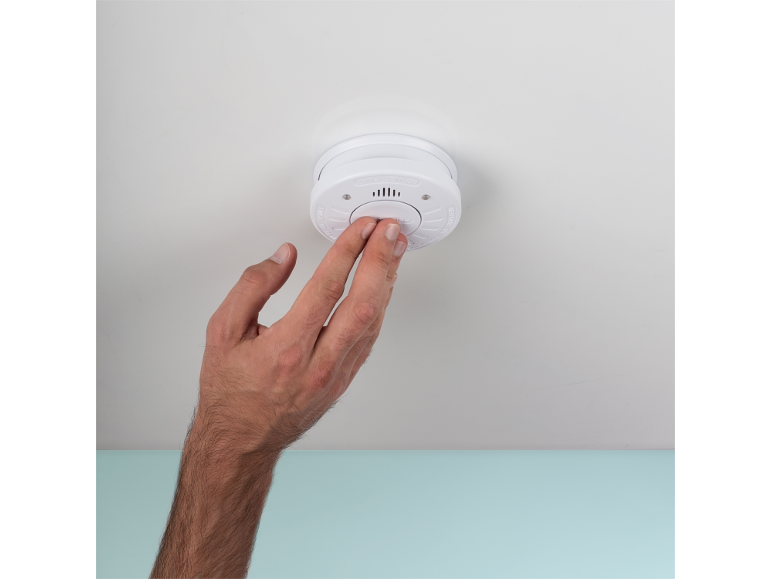 Una mano preme il pulsante di prova su un rilevatore di fumo montato a soffitto.