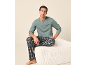 Uomo in maglia verde a maniche lunghe con scritta 'Slowdown' e pantaloni del pigiama a quadri, seduto su un pouf.