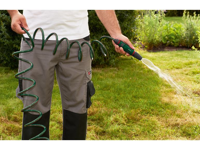 Uomo in pantaloni grigi innaffia il prato con un tubo a spirale verde e una pistola a spruzzo.
