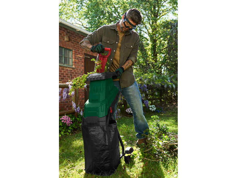 Uomo con cippatrice da giardino verde e rossa, che tritura rami in un giardino soleggiato.