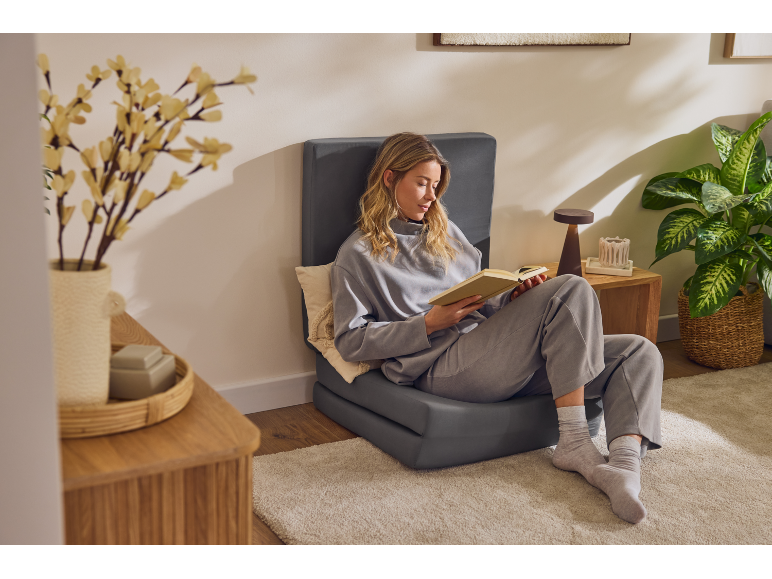 Donna che legge un libro su una seduta da terra grigia, con una pianta e un vaso di fiori.