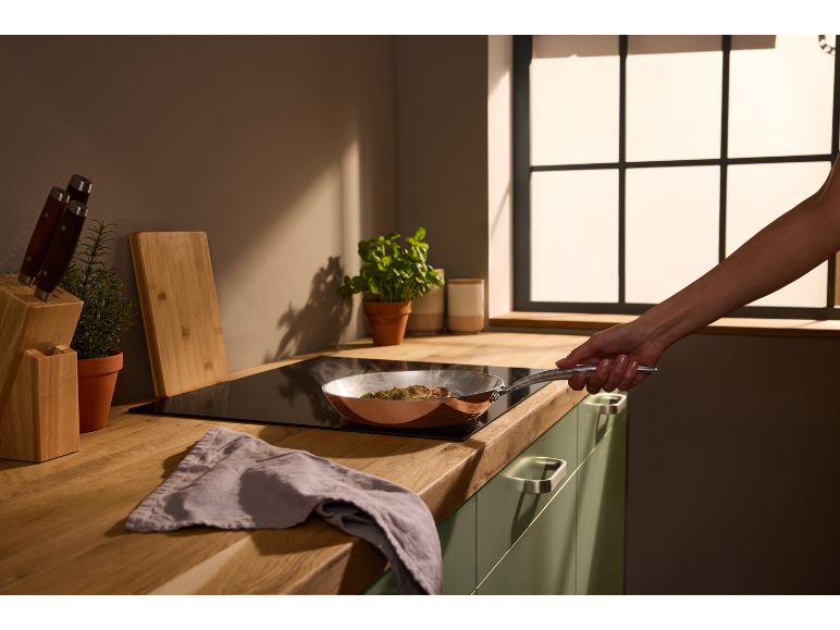 Cucina moderna con una padella di rame su un piano cottura, tenuta da una mano.