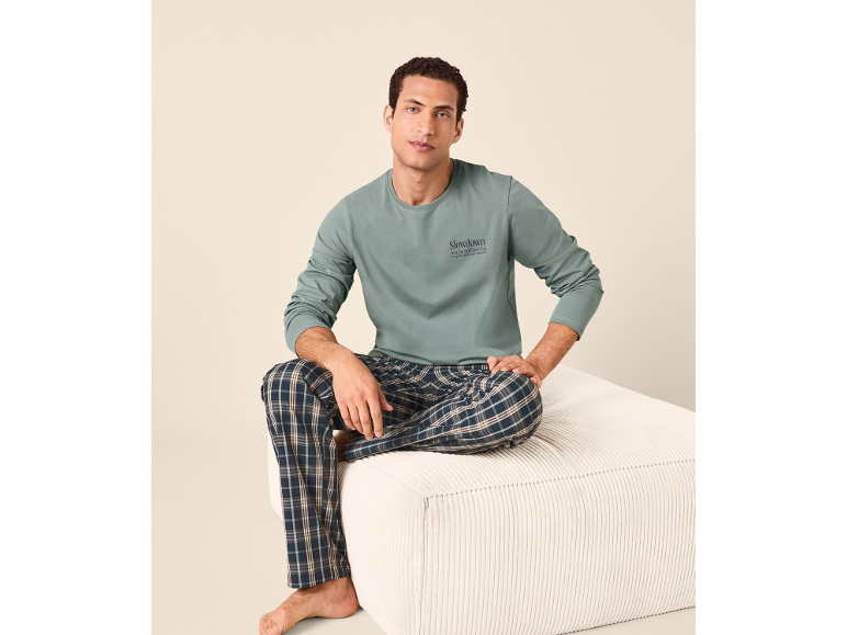 Uomo in maglia verde a maniche lunghe con scritta 'Slowdown' e pantaloni del pigiama a quadri, seduto su un pouf.
