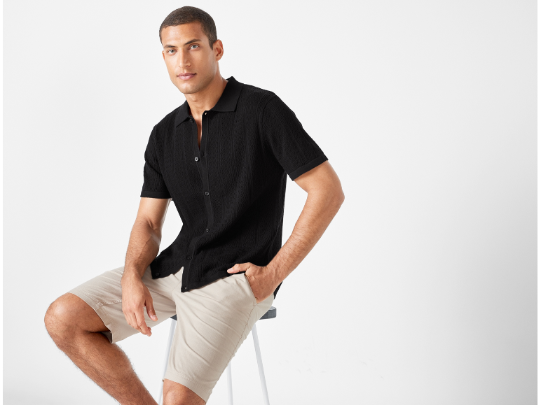 Uomo con camicia nera a maglia e pantaloncini beige, seduto su uno sgabello.