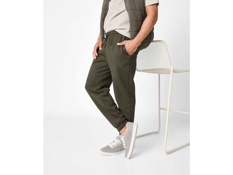 Uomo in joggers verde oliva, maglietta beige e gilet trapuntato, appoggiato a una sedia.