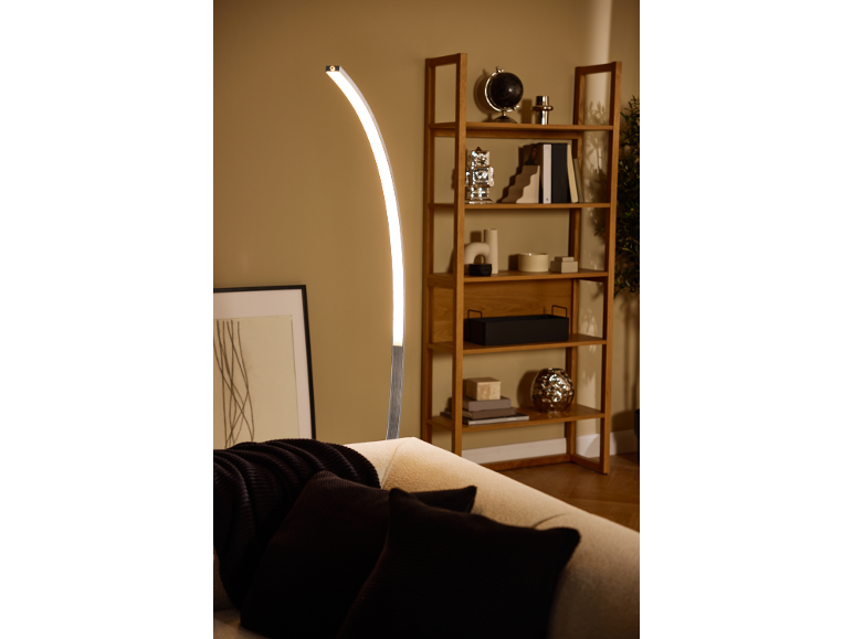 Lampada da terra LED curva illumina divano con cuscini e libreria in legno.