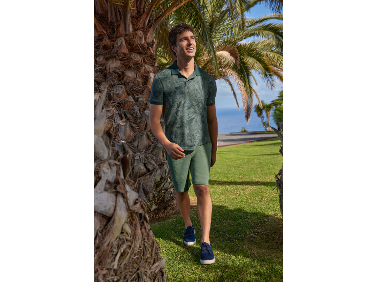 Uomo in polo verde fantasia e pantaloncini, cammina tra le palme in riva al mare.