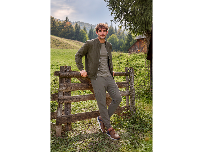 Uomo in giacca bomber color kaki, maglietta e pantaloni cargo, appoggiato a una staccionata di legno.