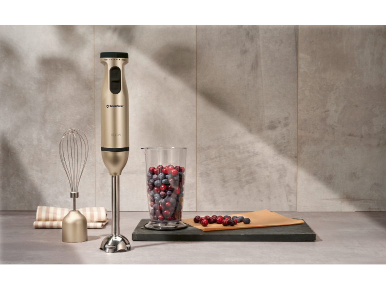 Frullatore a immersione dorato con frusta, misurino con frutti di bosco e strofinaccio.