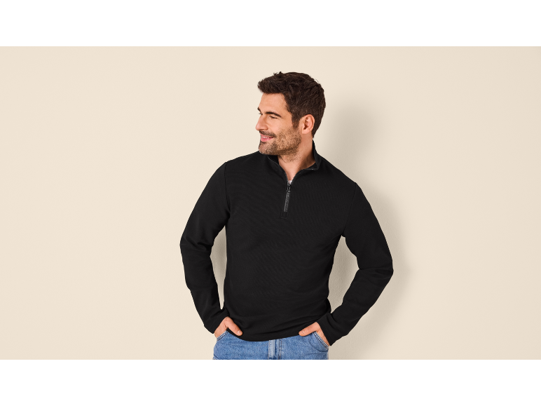Uomo in maglione nero con cerniera e jeans, mani nelle tasche.