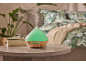 Diffusore di aromi verde con base in legno in una camera da letto con piante e biancheria da letto a fantasia.