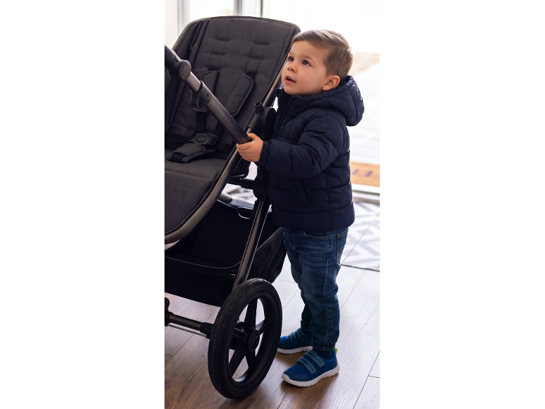 Bambino in giacca a vento blu navy e jeans, in piedi accanto a un passeggino grigio scuro.