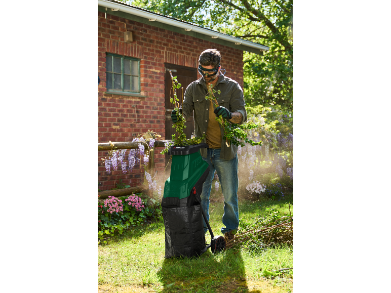 Uomo con occhiali di sicurezza e guanti usa un biotrituratore verde per rami.