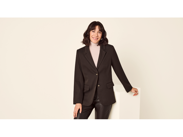 Donna con blazer nero, pantaloni in pelle e dolcevita beige, sorridente.