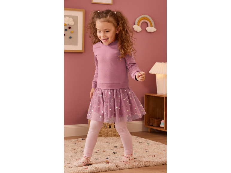 Bambina in felpa rosa, gonna in tulle e leggings, sorridente in una cameretta.