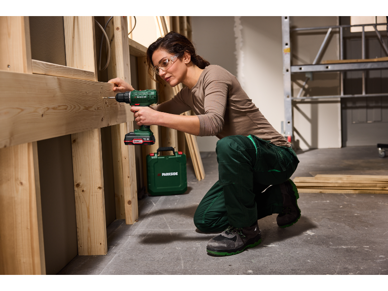 Donna con occhiali di sicurezza usa un trapano avvitatore a batteria su una struttura in legno.