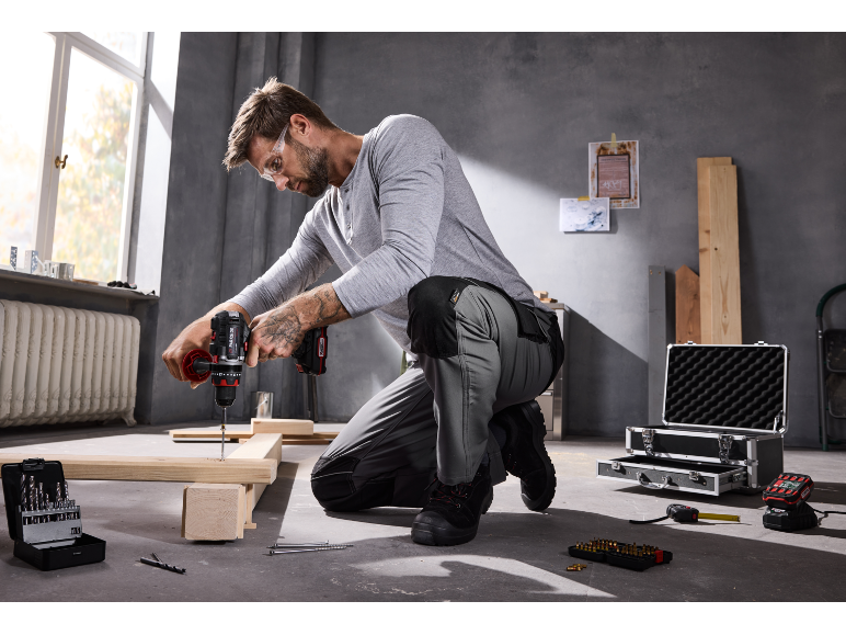 Uomo che fora il legno con un trapano a batteria, indossando occhiali di sicurezza e pantaloni da lavoro.