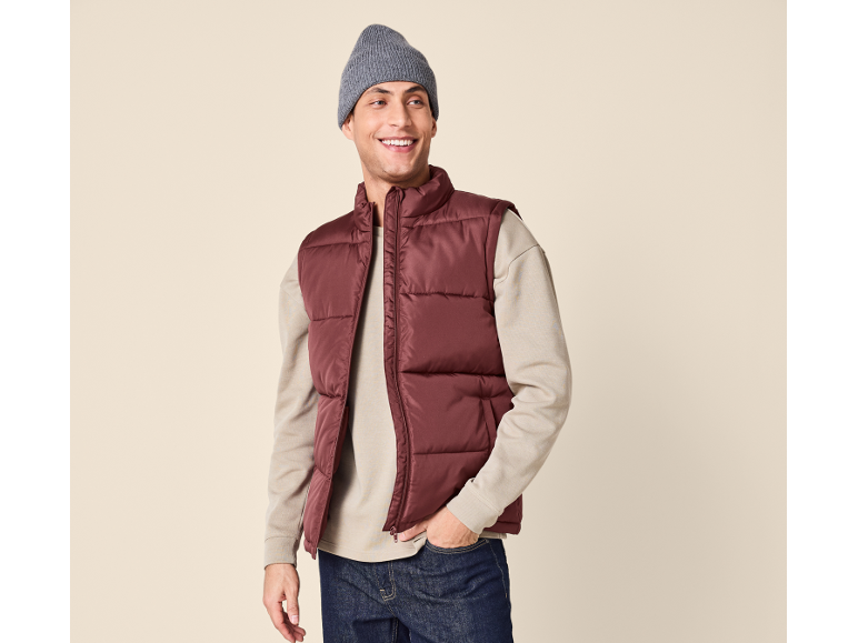 Uomo sorridente con gilet imbottito bordeaux, maglione beige e berretto grigio.