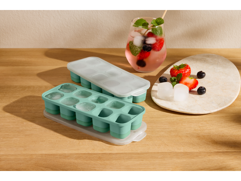 Due vassoi per cubetti di ghiaccio, uno pieno di ghiaccio, accanto a una bevanda ai frutti di bosco e frutta fresca.