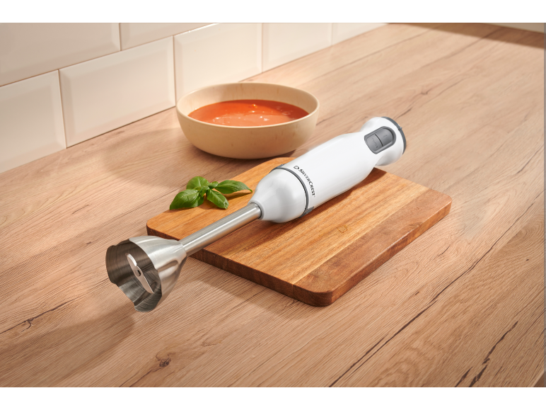Frullatore a immersione su un tagliere di legno accanto a una ciotola di zuppa di pomodoro e basilico.