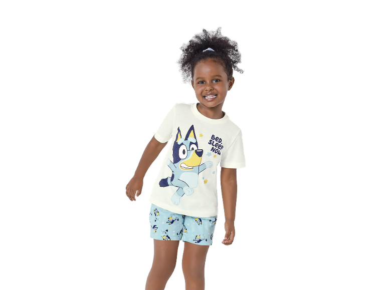 Bambina sorridente in pigiama bianco e azzurro con stampa di cane dei cartoni animati e scritta 'BED. SLEEP. NOW'.