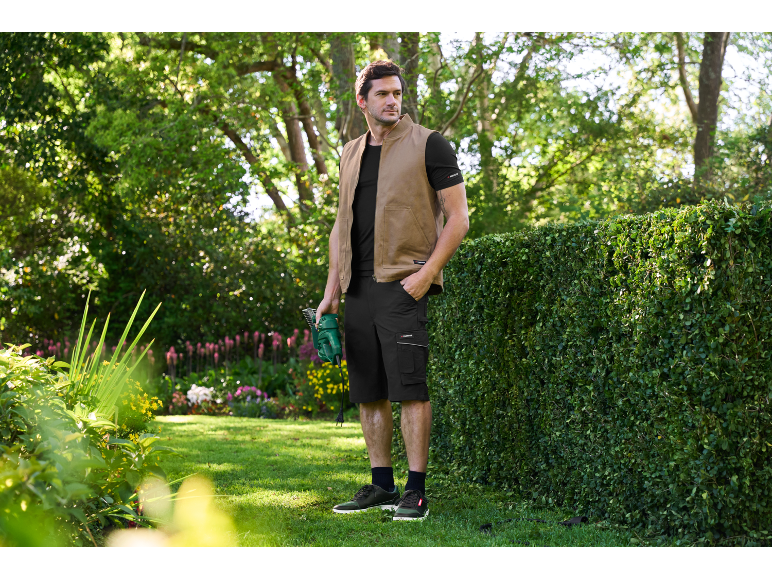 Uomo in gilet e pantaloncini che tiene un tagliasiepi in un giardino.