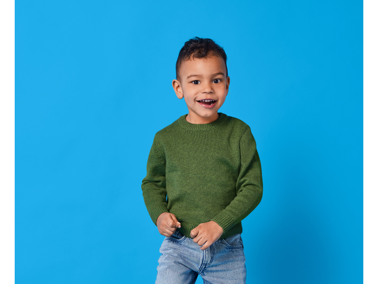 Bambino sorridente con maglione verde e jeans su sfondo blu.