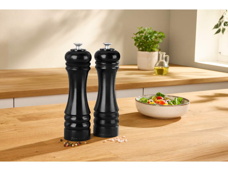 Due macinapepe neri su un bancone da cucina in legno con un'insalata fresca ed erbe aromatiche.