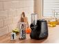 Robot da cucina nero con accessori per affettare e grattugiare, con verdure preparate.