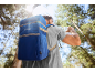Uomo con zaino frigo blu che beve acqua in una foresta soleggiata.