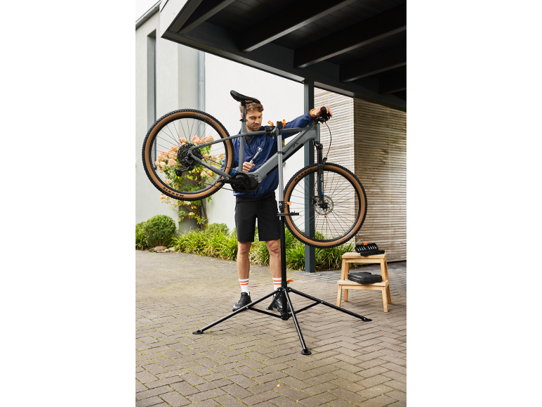 Uomo che ripara una bicicletta su un cavalletto da riparazione, con attrezzi su uno sgabello di legno.