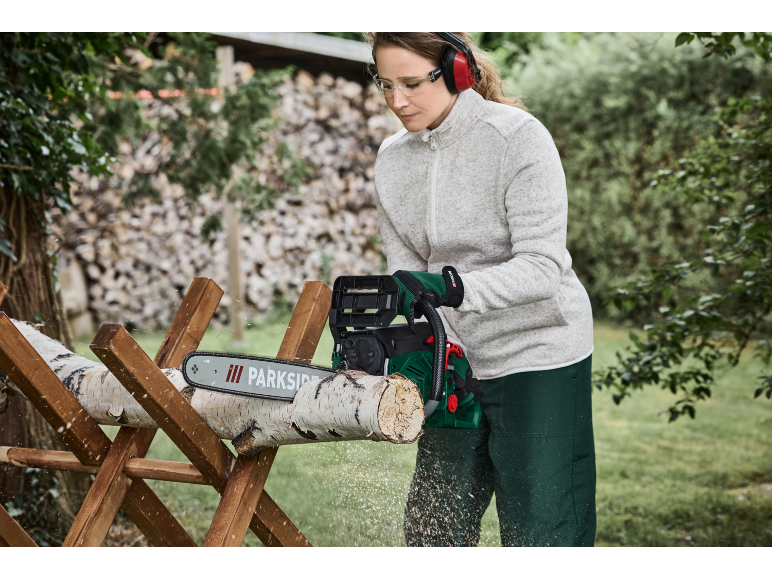 Donna con motosega che taglia un tronco di legno su un cavalletto, indossando protezioni per occhi e orecchie.