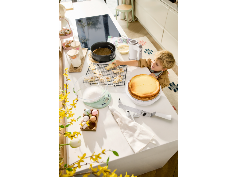 Bambino che prepara biscotti e torta in cucina, circondato da utensili e ingredienti.