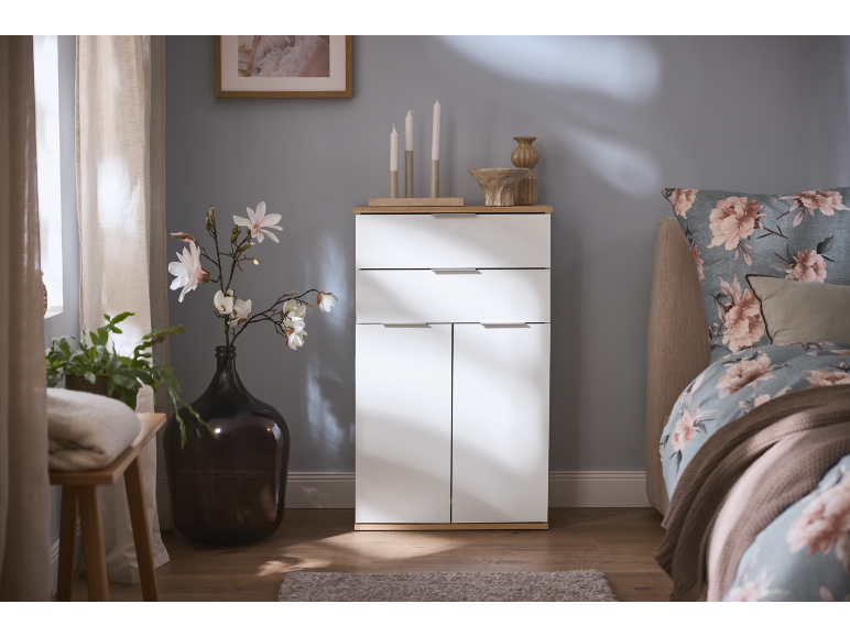 Mobile bianco con cassetti e ante, decorato con fiori e candele, accanto a un letto.