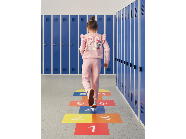 Bambina in tuta rosa con stampa di personaggio dei cartoni animati sulla schiena, cammina in un corridoio con armadietti blu.