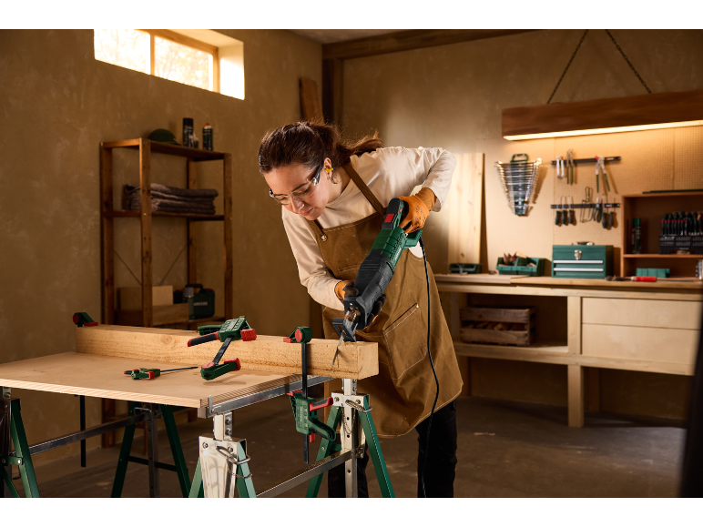 Donna con occhiali di sicurezza e guanti taglia legno con un seghetto alternativo in officina.