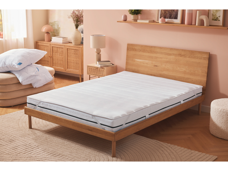 Camera da letto con materasso bianco, cuscini e piumino su un pouf, e mobili in legno.