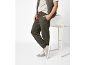 Uomo in joggers verde oliva, maglietta beige e gilet trapuntato, appoggiato a una sedia.