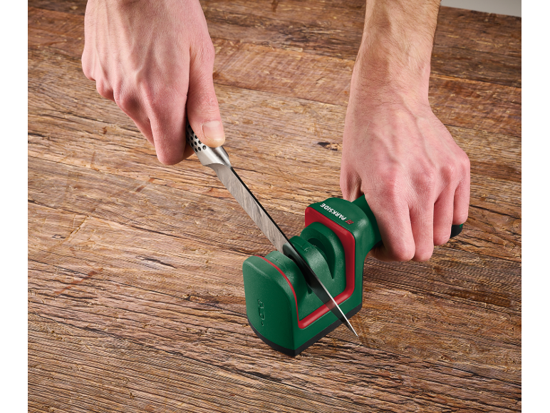 Uomo che affila un coltello con un affilacoltelli verde e rosso su un tavolo di legno.