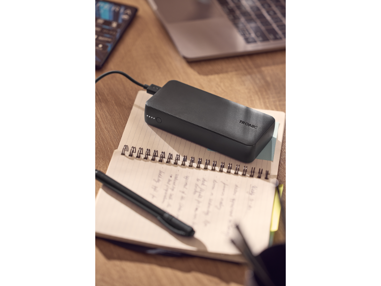 Power bank nero in carica su un quaderno aperto con una penna, un laptop sullo sfondo.